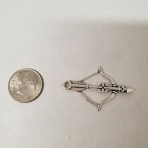 Op de afbeelding: Een zilverkleurige bedel in de vorm van een kruisboog met een pijl. De bedel ligt naast een Amerikaanse dime-munt. De kruisboogbedel is ongeveer 2,5 cm lang.