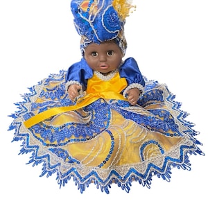 May include: A doll wearing a blue and yellow dress with a matching head wrap. The dress features intricate sequin and lace detailing. The doll has dark skin and is posed against a white background.