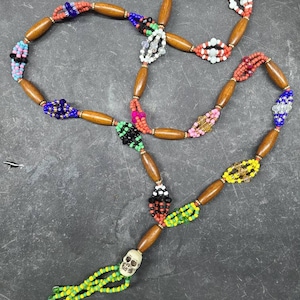 Peut inclure: Un collier de perles avec des perles en bois et des perles de verre colorées dans divers motifs. Le collier a un pendentif en forme de crâne et une gland de perles vertes et jaunes. Les perles sont disposées de manière symétrique.