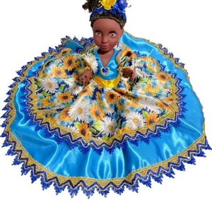 May include: A doll dressed in a blue and yellow dress with a sunflower pattern. The doll has dark skin and black hair with a yellow flower headband. The dress has a blue satin skirt with gold and blue trim.
