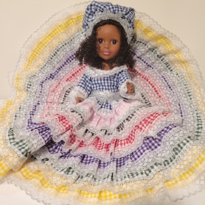 May include: A doll wearing a blue and white checkered dress with a pink and white ruffled skirt. The doll is wearing a blue and white checkered hat and has brown curly hair. The doll is sitting on a multi-colored skirt with white lace trim.