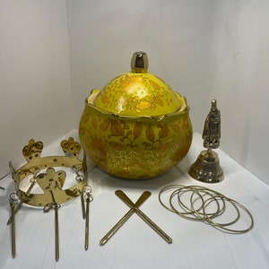 Può includere: Un vaso di ceramica giallo con un motivo floreale dorato e un coperchio dorato. Il vaso è circondato da oggetti in metallo dorato, tra cui una corona, una campana e un set di anelli.
