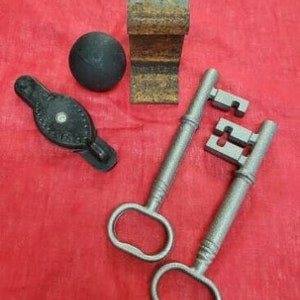 Peut inclure: Deux clés anciennes avec des motifs ornés, une petite boule noire et un bloc de métal rouillé. Les clés sont de couleur argentée et présentent des détails complexes. Le bloc est rectangulaire et a une texture rugueuse.