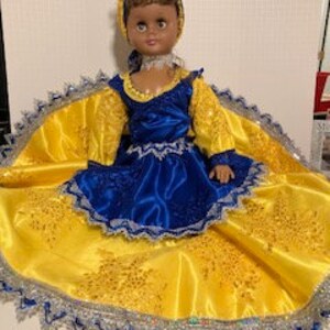 May include: A doll dressed in a blue and yellow outfit with a yellow head wrap. The doll has brown eyes and brown hair. The doll is wearing a blue dress with a yellow skirt and a yellow head wrap. The doll is sitting on a yellow and blue fabric.