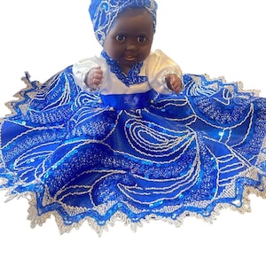 May include: A doll dressed in a blue and white outfit. The doll has dark skin and is wearing a blue head wrap. The dress is blue with silver accents and a white top. The doll is sitting down.