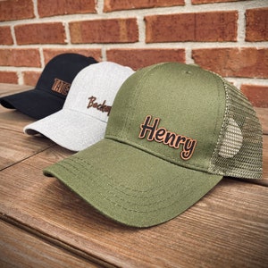 May include: Three baseball caps in a row, each with a different color and name. The first cap is black with the name "Tate" in a brown font. The second cap is gray with the name "Becker". The third cap is olive green with the name "Henry".