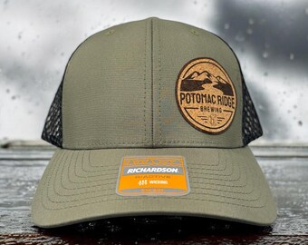 Puede incluir: Una gorra de camionero de malla verde y negra con un parche de cuero marrón que dice "Potomac Ridge Brewing". La gorra está hecha por Richardson y tiene una banda absorbente de humedad.