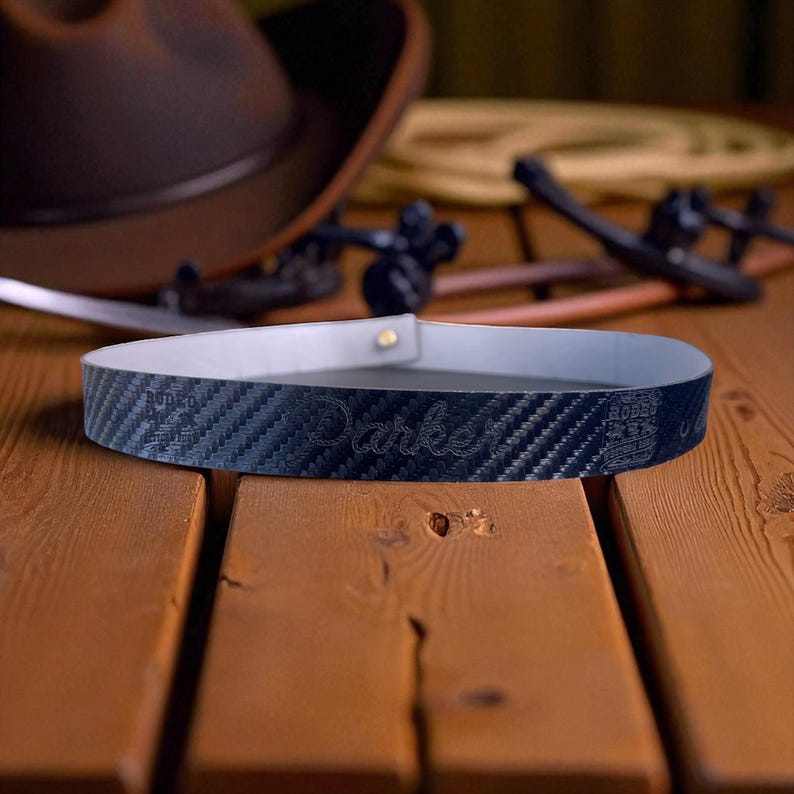 Pode incluir: Um cinto azul escuro com um padr&atilde;o de fibra de carbono e a palavra "Darke" bordada. O cinto tem um interior branco e um rebite dourado. Um chap&eacute;u de cowboy castanho e outros equipamentos de rodeio est&atilde;o no fundo.