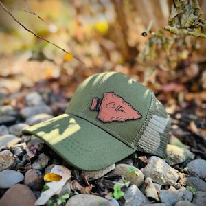 Puede incluir: Una gorra de béisbol verde y marrón con un parche de cuero marrón que dice "Colton". El parche tiene forma de punta de flecha.