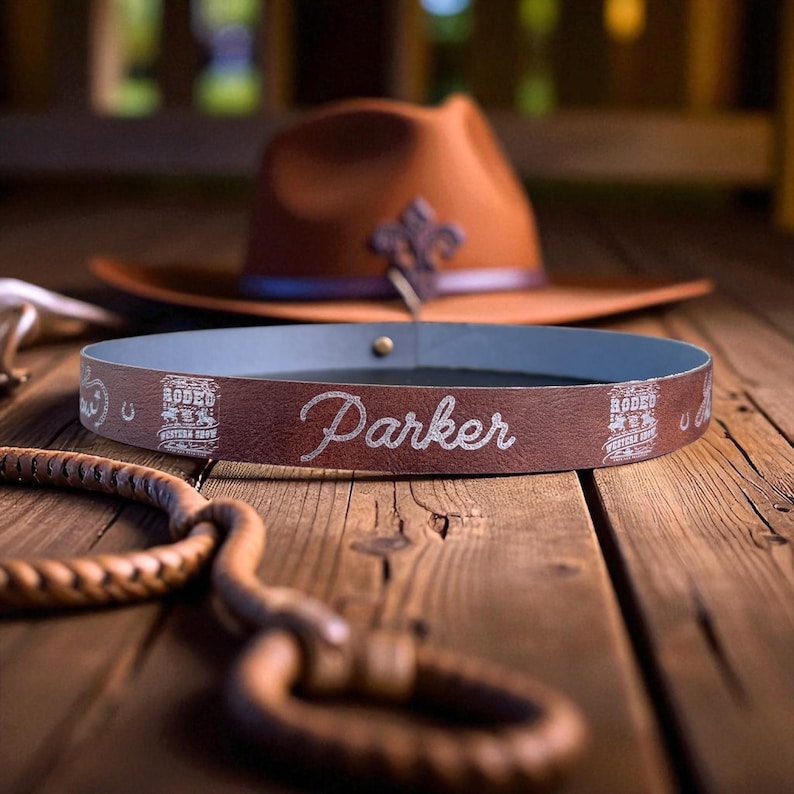Pode incluir: Uma faixa de chap&eacute;u de couro marrom com o nome "Parker" em escrita prateada. A faixa tem um interior azul e &eacute; decorada com gr&aacute;ficos tem&aacute;ticos de rodeio. Um chap&eacute;u de cowboy marrom e um chicote de couro tran&ccedil;ado est&atilde;o ao fundo.