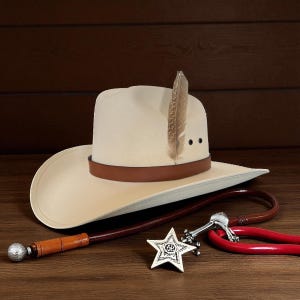 Può includere: Un cappello da cowboy color crema con una fascia in pelle marrone e una piuma. Sono presenti anche una frusta marrone, un distintivo argentato a forma di stella e un oggetto rosso a forma di ferro di cavallo. Gli oggetti sono disposti su una superficie di legno.
