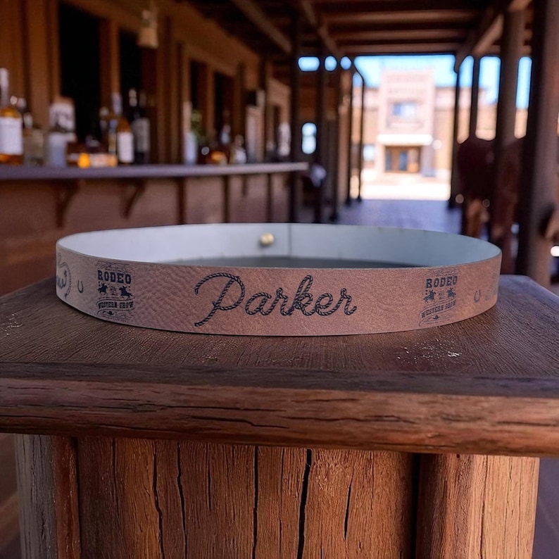 Pode incluir: Um cinto de couro castanho com o nome "Parker" em fonte cursiva. O cinto tem um interior branco e o texto "Rodeo Western Crew" e um log&oacute;tipo na lateral. O cinto &eacute; exibido numa superf&iacute;cie de madeira.