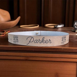 Pode incluir: Um cinto de couro bege claro com o nome "Parker" em fonte cursiva. O cinto tem um interior azul claro e &eacute; decorado com gr&aacute;ficos e texto com tema de rodeio. Um pequeno pino de metal prende o cinto.