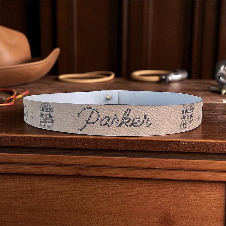 Pode incluir: Um cinto de couro bege claro com o nome "Parker" em fonte cursiva. O cinto tem um interior azul claro e &eacute; decorado com gr&aacute;ficos e texto com tema de rodeio. Um pequeno pino de metal prende o cinto.