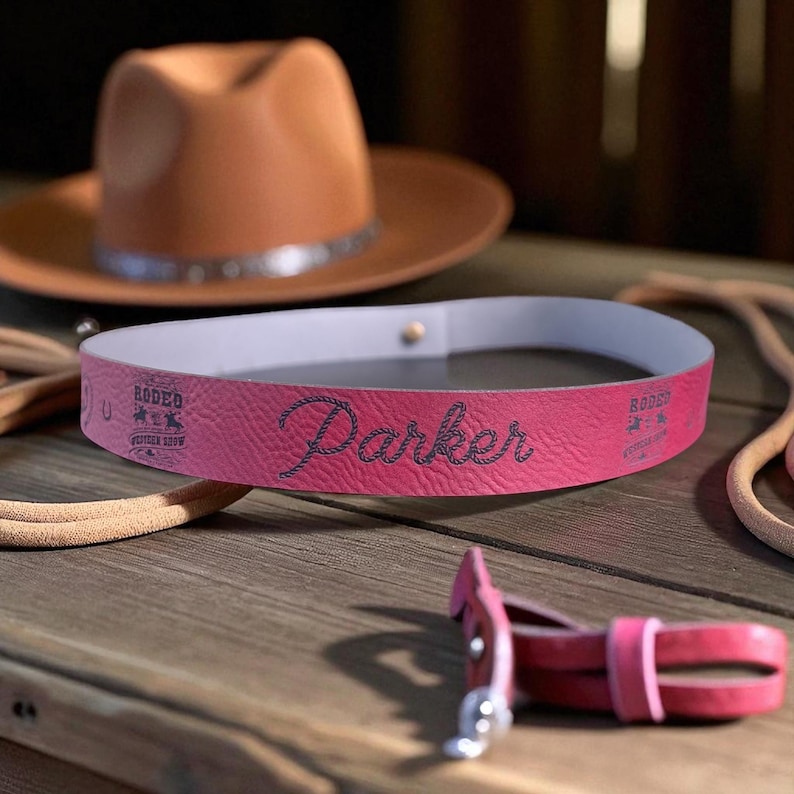 Pode incluir: Uma faixa de couro rosa com o nome "Parker" costurado em preto, juntamente com a palavra "Rodeo" e um logotipo. Um chap&eacute;u de cowboy marrom est&aacute; no fundo. Um cinto de couro rosa est&aacute; em primeiro plano.