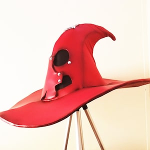 May include: A vibrant red witch hat with a wide brim and a unique, stylized face design. The hat features a pointed crown and is adorned with silver rivets. The hat is displayed on a tripod.