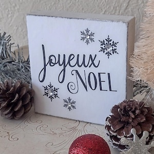 May include: A white wooden block with the words "Joyeux Noel" in black script, decorated with snowflake accents. The block is surrounded by pinecones, a red glitter ball, and a silver snowflake ornament, evoking a festive holiday theme.