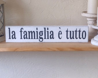 May include: A white wooden sign with the Italian phrase "la famiglia è tutto" in black lettering. The sign is on a wooden shelf, with a white lantern and two white candles on decorative holders.