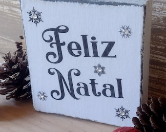 May include: A white wooden block with the words "Feliz Natal" in black script, decorated with snowflake accents. The block is surrounded by Christmas ornaments and pinecones, creating a festive holiday scene.