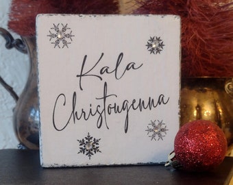 May include: A white square wooden sign with the words "Kala Christougenna" in black script, decorated with snowflake designs and rhinestones. A red glitter bauble sits nearby. The sign is placed on a stack of books.