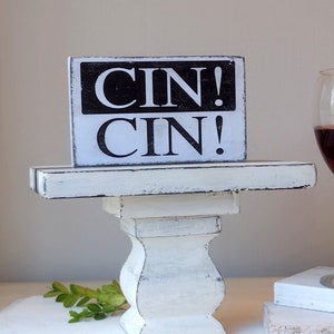 May include: White wooden pedestal cake stand with a black and white sign that reads "CIN! CIN!" on top. The stand has a distressed white finish and a pedestal base. A glass of red wine is in the background.
