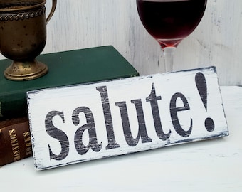May include: White wooden sign with black painted text that reads "salute!" The sign is distressed and has a rustic look.