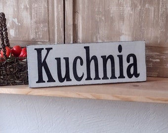 May include: A white wooden sign with black lettering that reads "Kuchnia" which is Polish for "Kitchen".