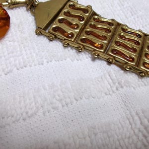 May include: A gold-toned chain watch fob with an amber-colored glass pendant. The fob has a decorative design of repeating curved lines.