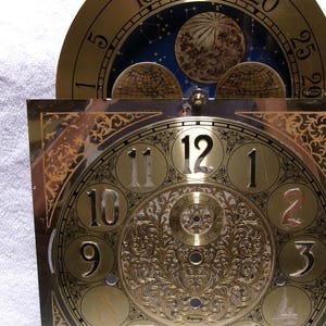 May include: A brass clock face with Roman numerals and ornate detailing. The clock face features a circular top section with celestial imagery and a lower square section with the numbers 1 through 12. The clock face is set against a white background.