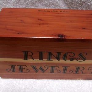 May include: A rectangular wooden jewelry box with a reddish-brown finish. The box is labeled "RINGS JEWELRY" in black lettering. The top of the box has the words "Ruidoso, New Mexico" in a smaller font.