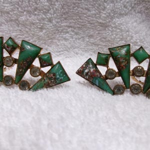 May include: A pair of vintage earrings with a geometric design. Each earring features green triangular and square-shaped stones, accented with small, clear, round stones. The earrings have a gold-colored metal frame.