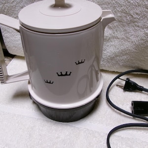 May include: Off-white electric coffee pot with a black base and handle. The pot has a lid with a knob and three black crown designs. A black power cord is coiled next to the pot.
