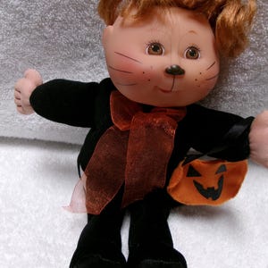 May include: A Halloween-themed doll with a cat-like face, wearing a black outfit and holding an orange pumpkin-shaped bag. The doll has red hair, a brown ribbon, and painted whiskers. The doll is approximately 8 inches tall.