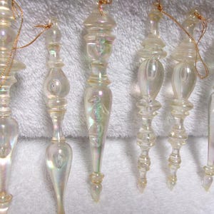 May include: Six iridescent glass Christmas ornaments with gold-colored string hangers. The ornaments have a variety of shapes, including teardrop and elongated designs. The clear glass reflects light, creating a shimmering effect, perfect for holiday decor.