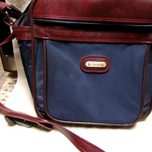 May include: A navy blue Samsonite shoulder bag with burgundy leather trim and a matching adjustable strap. The bag features a zippered top closure and a small rectangular logo on the front. The bag is sitting on a white towel.