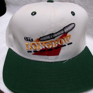May include: White and green baseball cap with the text "GTE LONGBOW" embroidered on the front. The cap features a dark green brim and a design of a silver object with a red and yellow background.