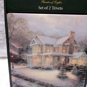 May include: Set of 2 trivets featuring a winter scene of a snow-covered house with lit windows and a snowman. The trivets are packaged in a dark green box with the text "Thomas Kinkade Painter of Light".