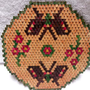 May include: A woven trivet with a geometric design. The trivet is hexagonal with a light brown background and a dark brown, green, and red floral pattern. The border is green.