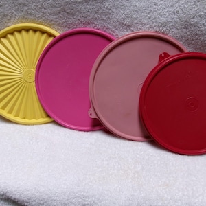 May include: Four colorful Tupperware lids in yellow, pink, light pink, and red. The yellow lid has a sunburst design. The lids are arranged on a white towel.