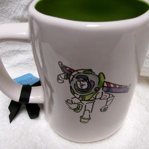 May include: A white ceramic mug with a green interior featuring a stylized line-art illustration of the character Buzz Lightyear in his space suit. A black ribbon is tied around the handle, and a small blue tag is attached to the handle.