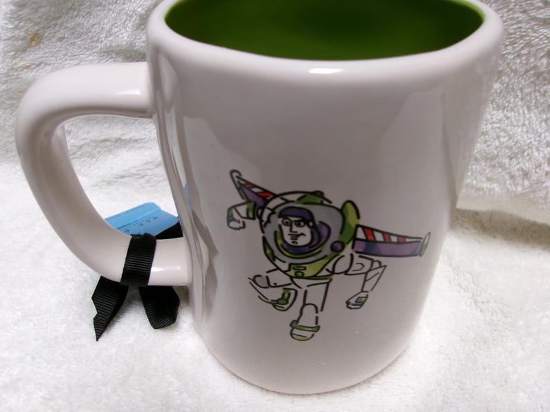 May include: A white ceramic mug with a green interior featuring a stylized line-art illustration of the character Buzz Lightyear in his space suit. A black ribbon is tied around the handle, and a small blue tag is attached to the handle.
