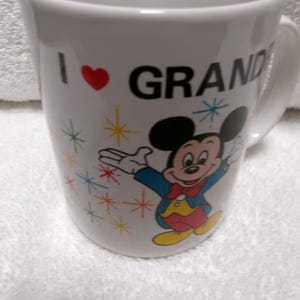 Op de afbeelding: Witte keramische mok met de tekst "I ❤️ GRAND" en een cartoonafbeelding van Mickey Mouse. De mok heeft een rood hart en kleurrijke sterrenpatronen. Het handvat is wit.
