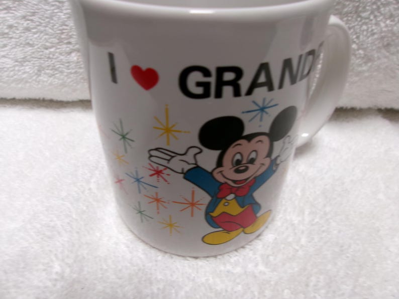 Op de afbeelding: Witte keramische mok met de tekst "I ❤️ GRAND" en een cartoonafbeelding van Mickey Mouse. De mok heeft een rood hart en kleurrijke sterrenpatronen. Het handvat is wit.