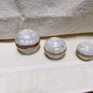 May include: Three round, decorative boxes made of white mother of pearl with gold trim. The boxes are arranged in a row from largest to smallest.