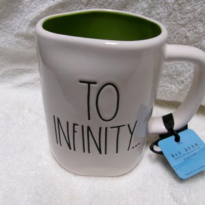 May include: A white ceramic mug with a lime green interior and a handle on the right side. The exterior features the black text TO INFINITY... in a handwritten style. A blue rectangular product tag is attached to the handle with a black cord.