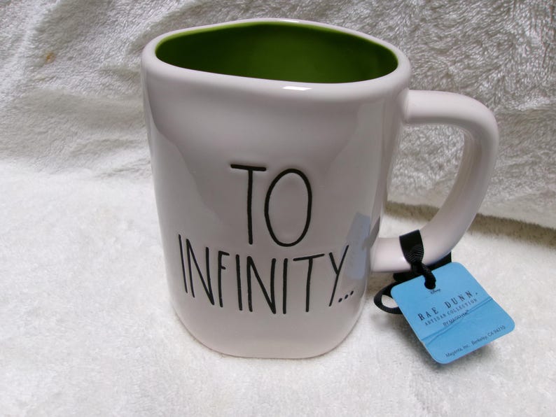 May include: A white ceramic mug with a lime green interior and a handle on the right side. The exterior features the black text TO INFINITY... in a handwritten style. A blue rectangular product tag is attached to the handle with a black cord.