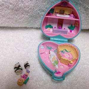 May include: A vintage heart-shaped Polly Pocket playset in pink and teal, open to reveal a miniature interior with furniture and accessories. Includes small panda figures and a miniature person figure.