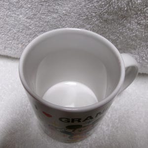 Op de afbeelding: Witte keramische mok met een handvat. De mok heeft een kleurrijke afbeelding van Mickey Mouse en het woord "GRANDMA". De mok heeft een glanzende afwerking en wordt waarschijnlijk gebruikt voor het drinken van dranken.