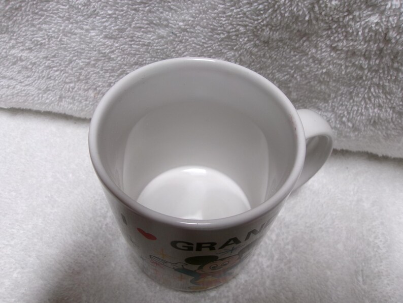 Op de afbeelding: Witte keramische mok met een handvat. De mok heeft een kleurrijke afbeelding van Mickey Mouse en het woord "GRANDMA". De mok heeft een glanzende afwerking en wordt waarschijnlijk gebruikt voor het drinken van dranken.