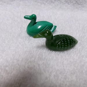 May include: Two decorative duck figurines. One is a solid, glossy green, and the other is a darker green with white dot details. Both are positioned on a white, textured surface. The ducks are likely made of a smooth, polished material.